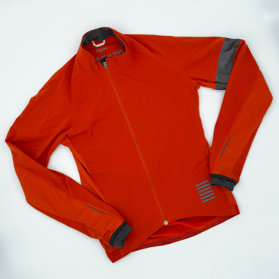 Rapha Pro Team Jacket angled front showing rust softshell, offset zip and charcoal armband